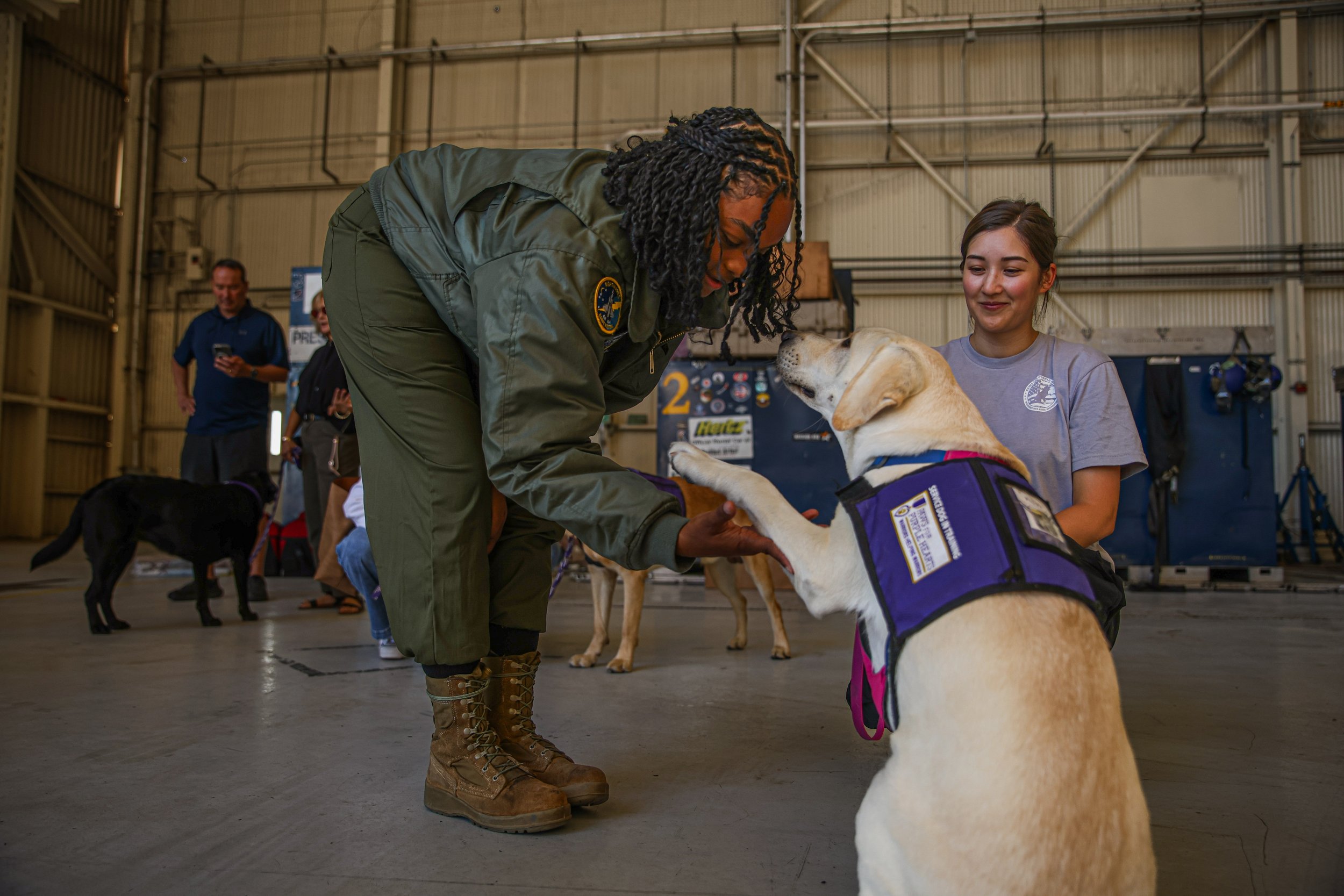 A man wearing a cream knit beanie and ripped black jeans sitting in a chair while hugging a black Labrador dog. The man is smiling and the dog is lying across his lap. The setting appears to be indoors with a white wall and some chairs and a table in the background.